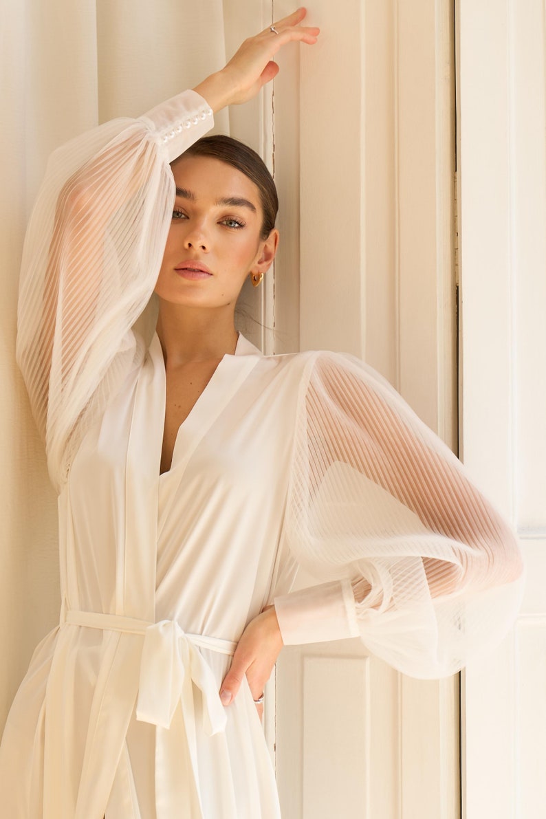 May include: A white robe with sheer, pleated sleeves and a sash tie. The robe has a V-neckline and a relaxed fit.