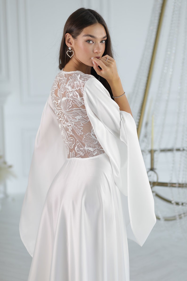 May include: A white satin dress with a lace back and long sleeves. The dress has a flowing skirt and a fitted bodice.