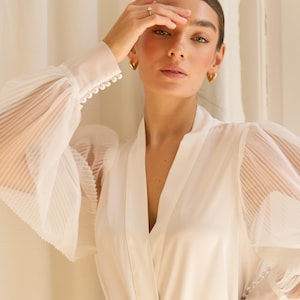 May include: A white satin robe with sheer, pleated sleeves and pearl detailing. The robe has a V-neckline and a tie belt.