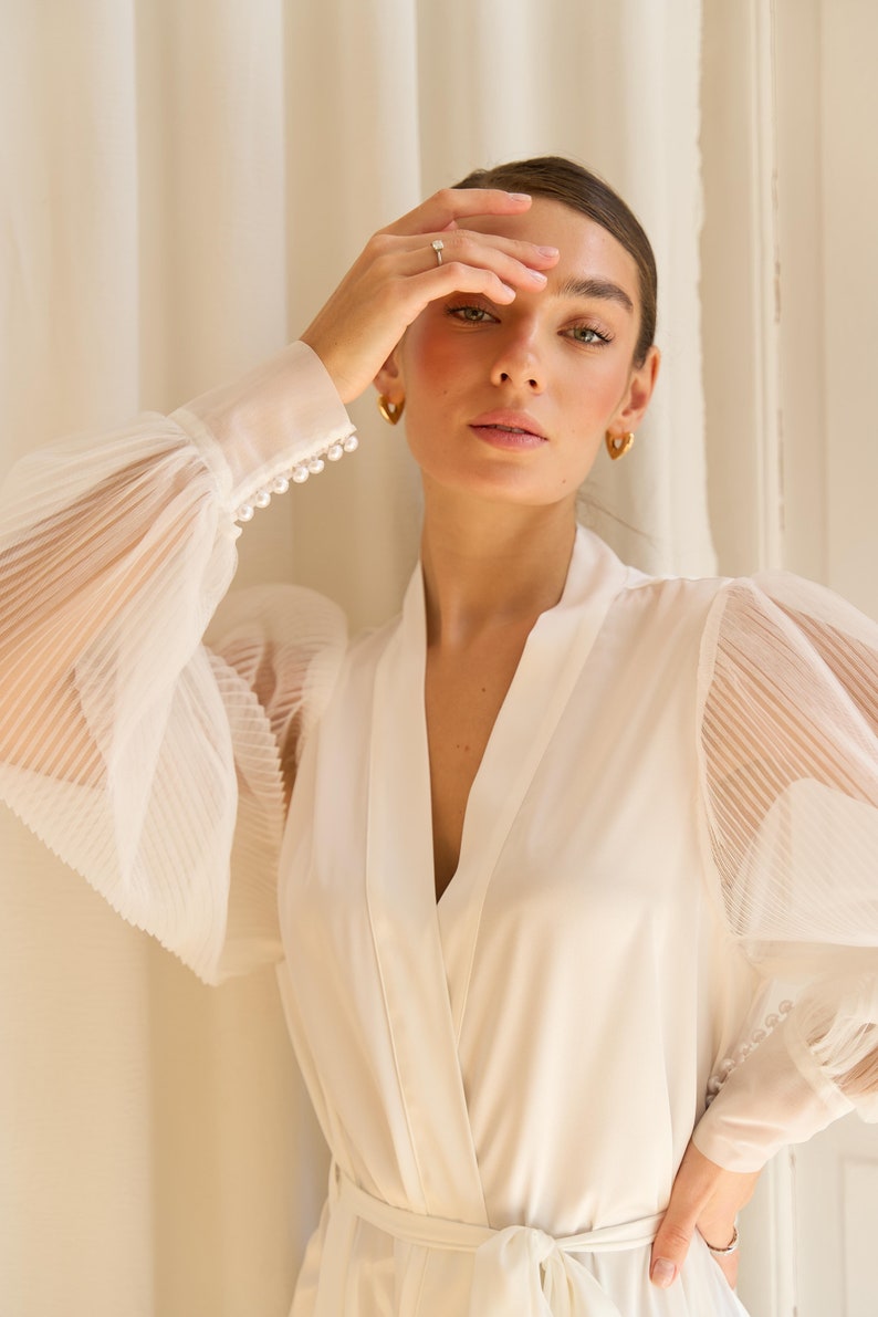 May include: A white satin robe with sheer, pleated sleeves and pearl detailing. The robe has a V-neckline and a tie belt.
