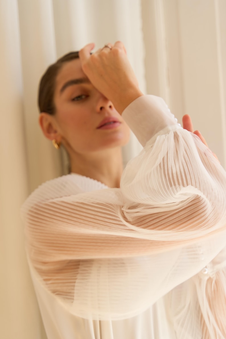 May include: A woman wearing a white sheer blouse with pleated detailing. The blouse has long sleeves with puffed cuffs. The woman is looking down and has her hand on her face.