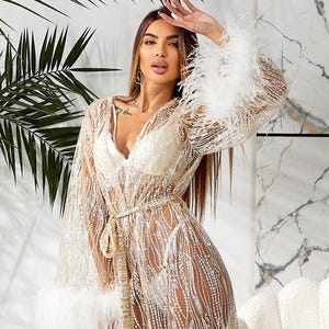 May include: A sheer, gold-colored robe with a tie waist and white feather trim on the sleeves. The robe is embellished with sparkling details and has a V-neck design. The garment is part of a lingerie or loungewear collection.