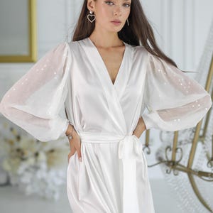 May include: A white ceramic mug with a black handle and the text "But First, Coffee" in a bold, black font. The mug is set against a blurred background, suggesting a cosy setting.