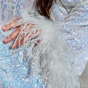 May include: A long-sleeved garment covered in iridescent, light-reflecting sequins that shift between shades of white, silver, and pale blue. The sleeve cuff is finished with a thick, voluminous trim of long, wispy white ostrich feathers.
