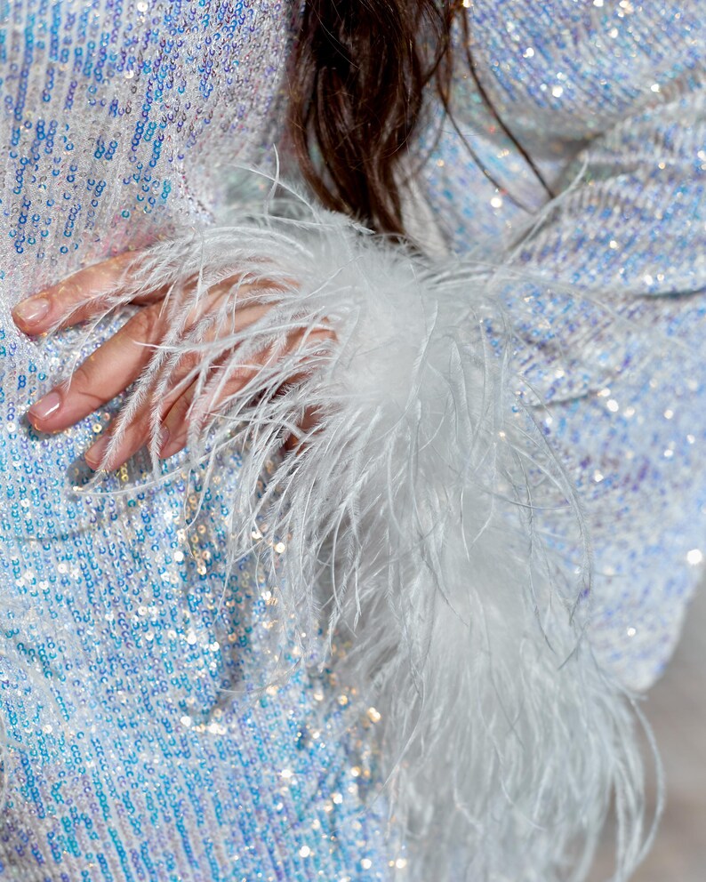 May include: A long-sleeved garment covered in iridescent, light-reflecting sequins that shift between shades of white, silver, and pale blue. The sleeve cuff is finished with a thick, voluminous trim of long, wispy white ostrich feathers.