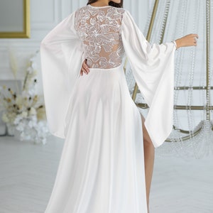 May include: A white maxi dress with a floral lace back detail and long, flowing sleeves. The dress has a high neckline and a slit up the front.