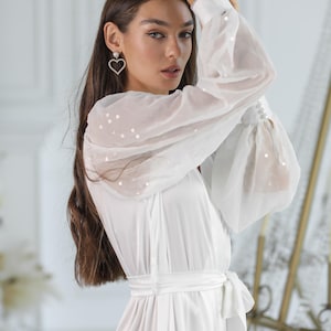 May include: A white satin robe with sheer, sequined sleeves and a matching tie belt. The robe features a V-neckline and is paired with heart-shaped earrings. The sleeves have a gathered cuff detail.
