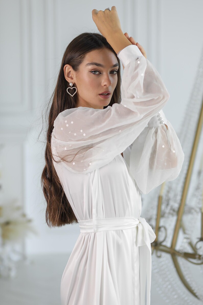 May include: A white satin robe with sheer, sequined sleeves and a matching tie belt. The robe features a V-neckline and is paired with heart-shaped earrings. The sleeves have a gathered cuff detail.