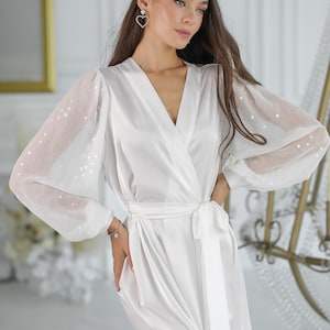 May include: A white satin robe with a wrap design and a matching tie belt. The sleeves are sheer with a polka dot pattern. The robe has a V-neck and a thigh-high slit. The model is wearing heart-shaped earrings.