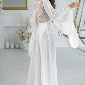 May include: A white satin gown with long sleeves and a lace back. The sleeves are wide and flowy, and the gown has a flowing skirt.