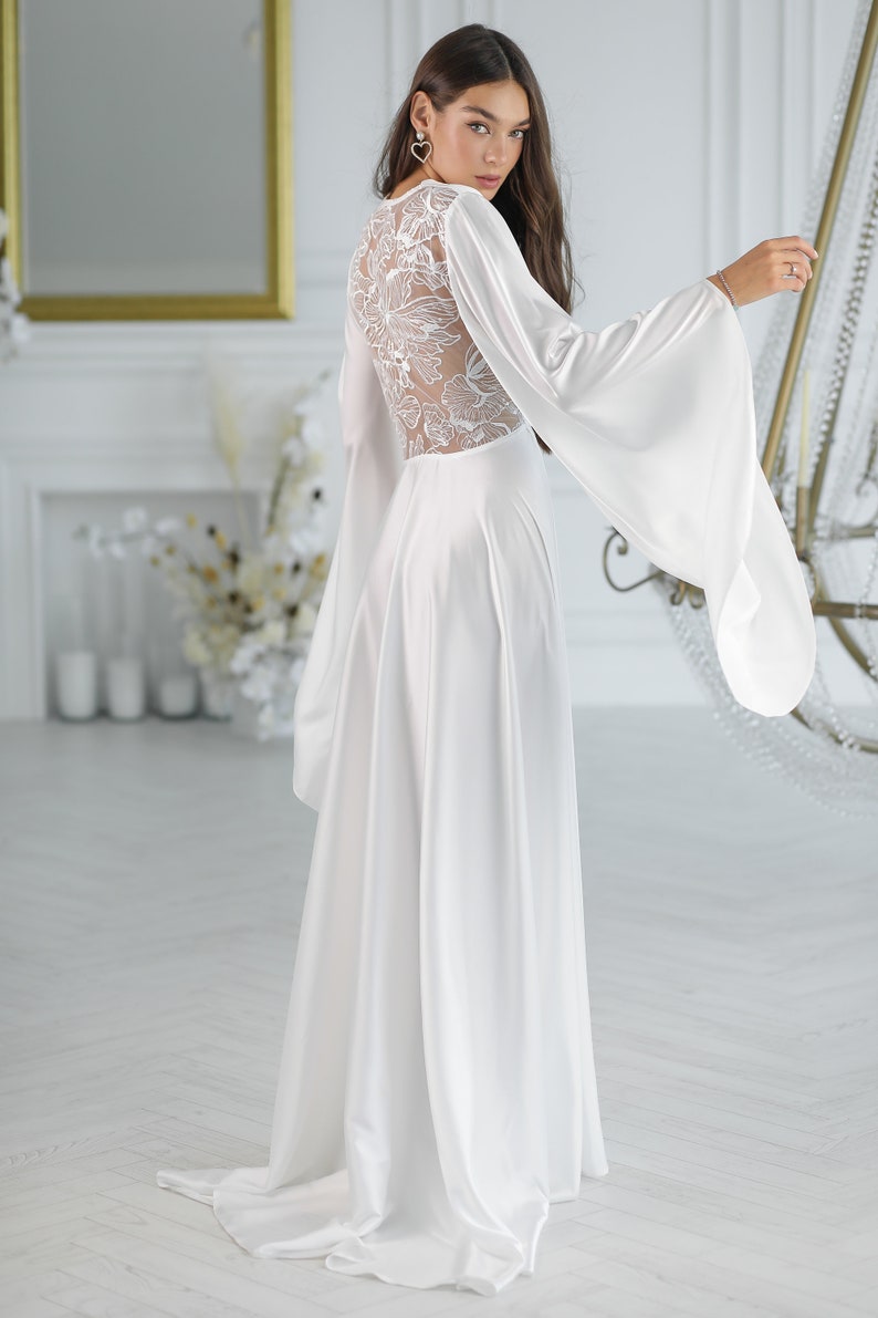 May include: A white satin gown with long sleeves and a lace back. The sleeves are wide and flowy, and the gown has a flowing skirt.
