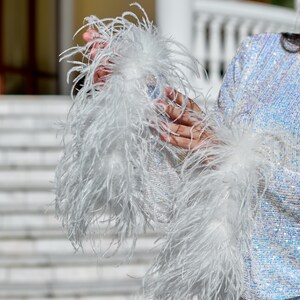 May include: A long-sleeved top covered in iridescent, light-reflecting sequins. The sleeves feature prominent, voluminous white ostrich feather cuffs that extend from the wrists, creating a textured and layered appearance against the shimmering fabric.