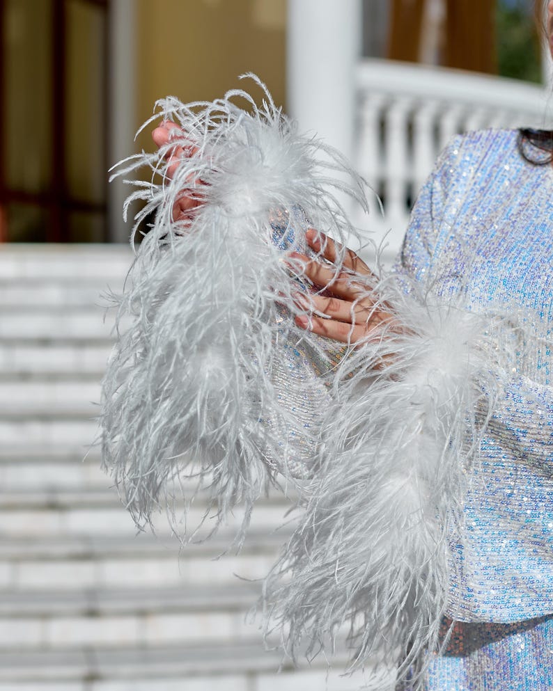 May include: A long-sleeved top covered in iridescent, light-reflecting sequins. The sleeves feature prominent, voluminous white ostrich feather cuffs that extend from the wrists, creating a textured and layered appearance against the shimmering fabric.