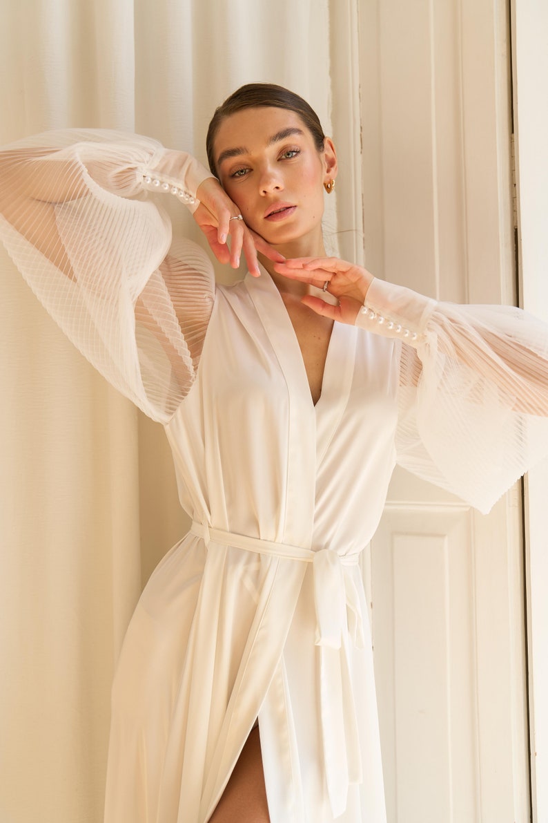 May include: A white satin robe with long sleeves and a tie belt. The sleeves are sheer and have a pleated design. The robe has pearl buttons on the cuffs.