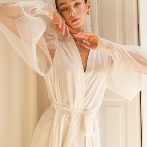 May include: A white satin robe with long sleeves and a tie belt. The sleeves are sheer and have a pleated design. The robe has pearl buttons on the cuffs.