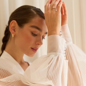 May include: A white sheer blouse with long, pleated sleeves and pearl button detailing on the cuffs.