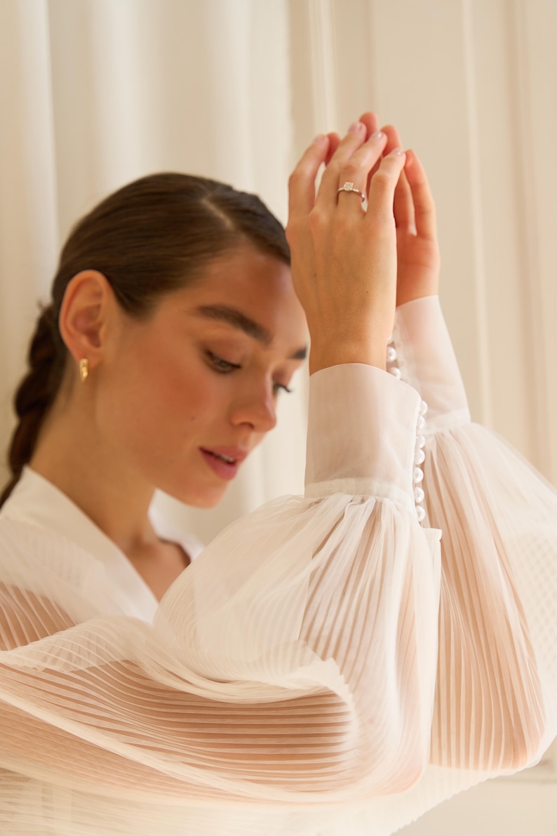 May include: A white sheer blouse with long, pleated sleeves and pearl button detailing on the cuffs.