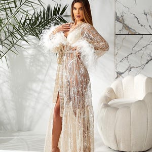 May include: A long, flowing, gold sequined robe with a high slit and feather trim on the sleeves. The robe has a V-neck and is paired with nude heels. The background includes a white chair and palm fronds.