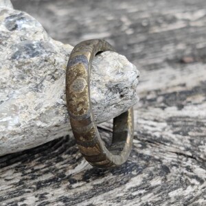 Op de afbeelding: Een bronzen ring met een getextureerd oppervlak, rustend op een grijze steen. Het ontwerp van de ring heeft een herhalend patroon van kleine, cirkelvormige vormen. De achtergrond is een verweerd houten oppervlak.