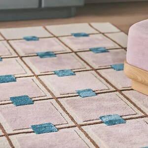 Op de afbeelding: Een rechthoekig tapijt met een rasterpatroon. Het tapijt heeft vierkanten in roze en beige tinten, omlijnd met bruine lijnen. Turquoiseblauwe vierkanten zijn in het midden van sommige roze vierkanten geplaatst. Een roze poef met houten poten staat op de achtergrond.