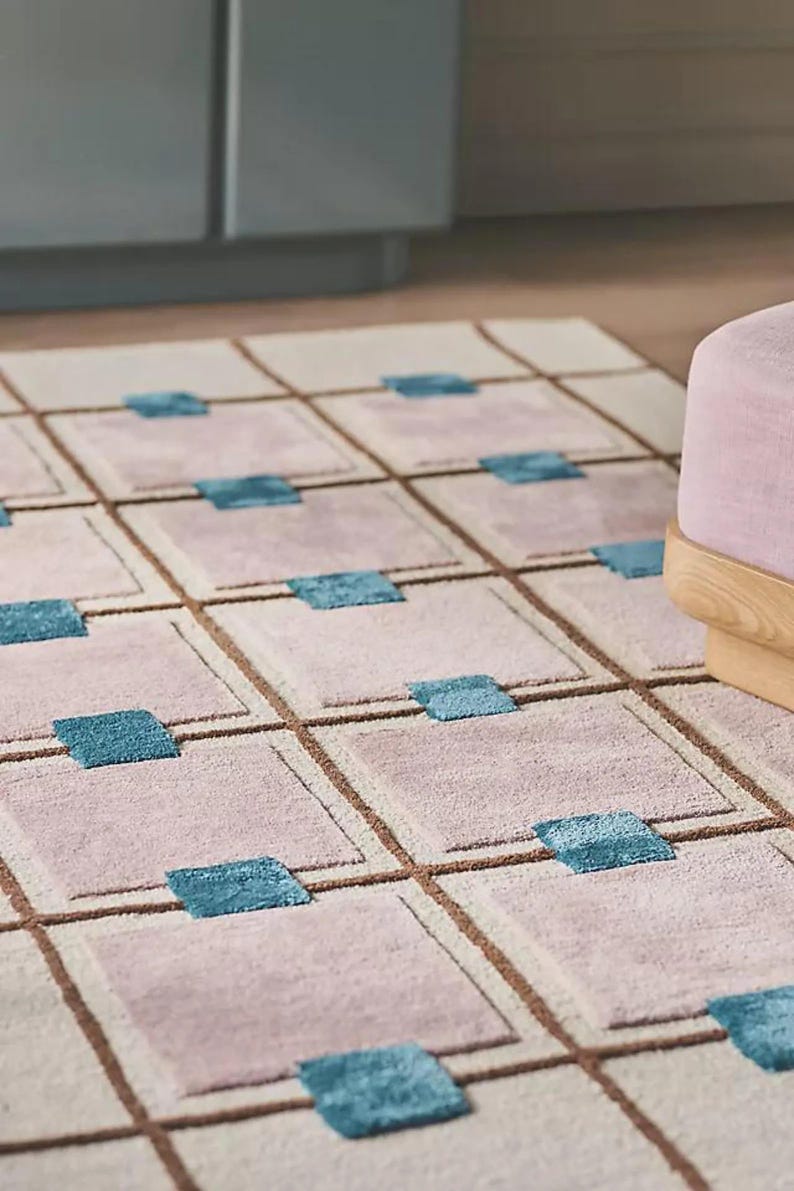 Op de afbeelding: Een rechthoekig tapijt met een rasterpatroon. Het tapijt heeft vierkanten in roze en beige tinten, omlijnd met bruine lijnen. Turquoiseblauwe vierkanten zijn in het midden van sommige roze vierkanten geplaatst. Een roze poef met houten poten staat op de achtergrond.
