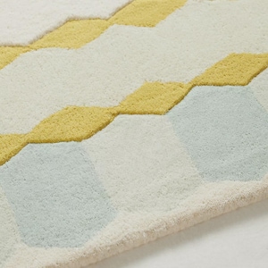 K&ouml;nnte beinhalten: Nahaufnahme eines weichen, strukturierten Teppichs mit geometrischem Muster. Der Teppich hat einen cremefarbenen Hintergrund mit gelben und hellblauen Rauten. Die R&auml;nder des Teppichs sind hellbeige.
