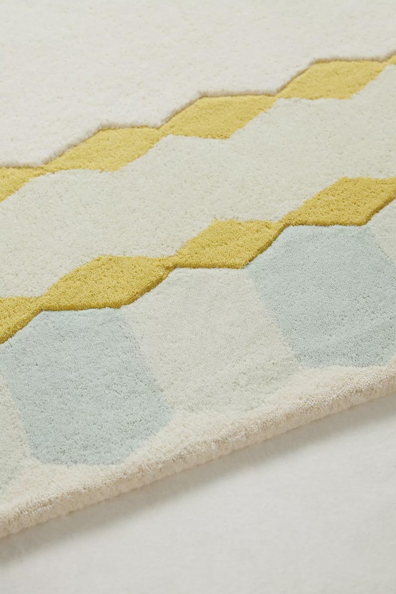 K&ouml;nnte beinhalten: Nahaufnahme eines weichen, strukturierten Teppichs mit geometrischem Muster. Der Teppich hat einen cremefarbenen Hintergrund mit gelben und hellblauen Rauten. Die R&auml;nder des Teppichs sind hellbeige.