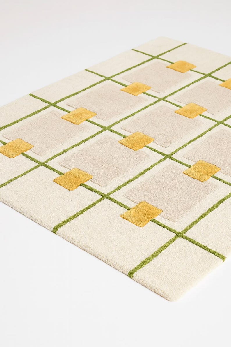 May include: Cream-colored rectangular rug with a geometric pattern. The design features a grid of green lines, beige squares, and yellow diamond accents. The rug has a textured surface and a modern aesthetic.