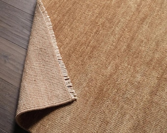 May include: A tan area rug with a textured, woven surface and a folded corner revealing a lighter beige, grid-patterned backing with a fringe edge. The rug features a consistent, dense pile construction throughout its rectangular surface area.