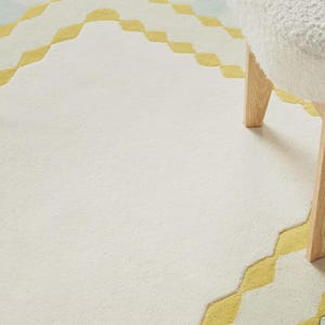 K&ouml;nnte beinhalten: Ein cremefarbener Teppich mit einem geometrischen Design in Gelb und Hellblau. Der Teppich hat eine weiche Textur und einen rautenf&ouml;rmigen Rand. Ein wei&szlig;er, flauschiger Hocker mit Holzbeinen ist teilweise sichtbar.