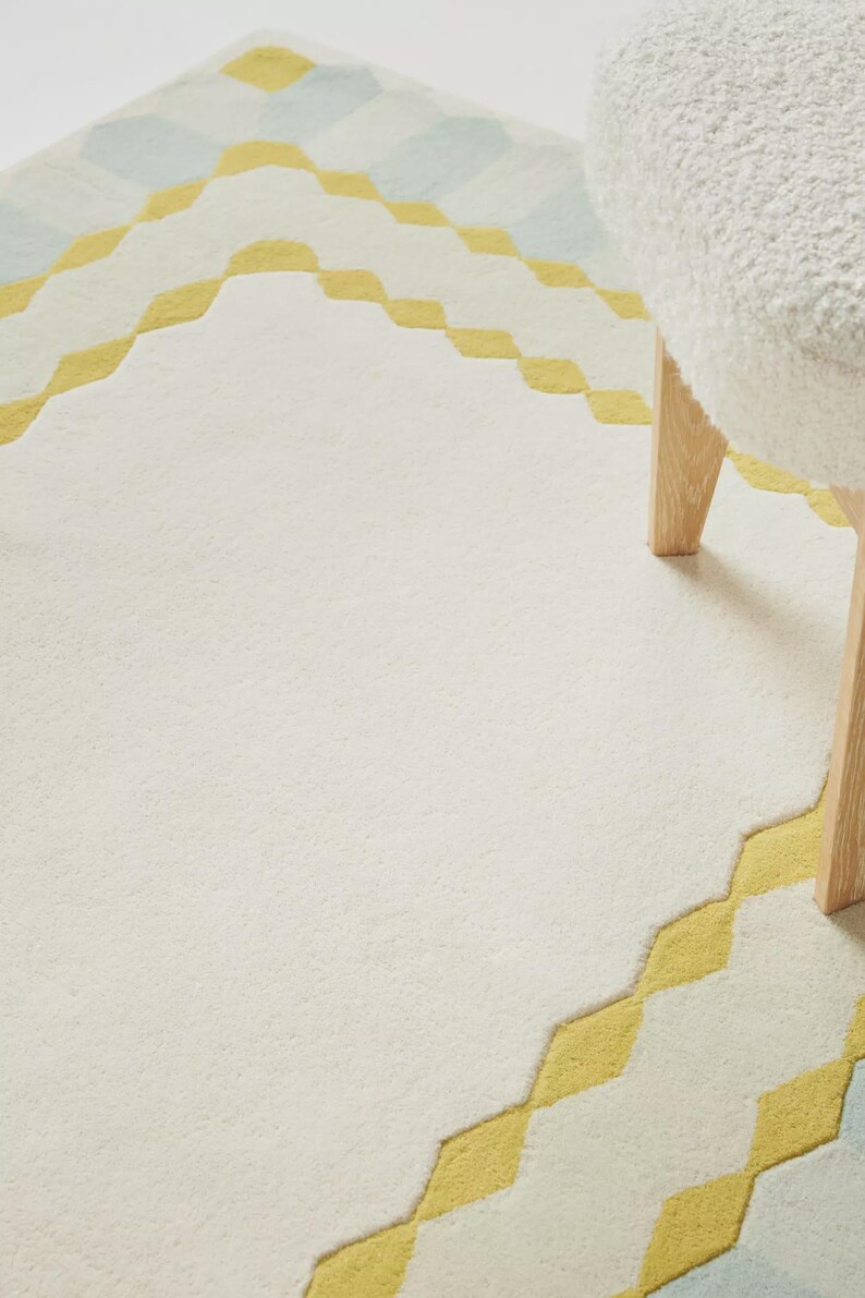K&ouml;nnte beinhalten: Ein cremefarbener Teppich mit einem geometrischen Design in Gelb und Hellblau. Der Teppich hat eine weiche Textur und einen rautenf&ouml;rmigen Rand. Ein wei&szlig;er, flauschiger Hocker mit Holzbeinen ist teilweise sichtbar.