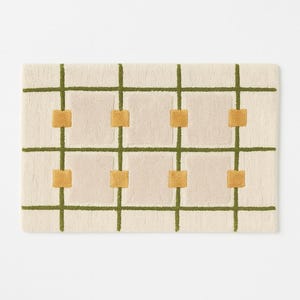 May include: Rectangular rug with a cream-colored background, featuring a grid pattern of olive green lines and squares. The squares are filled with a mustard yellow color. The rug has a soft, textured appearance.