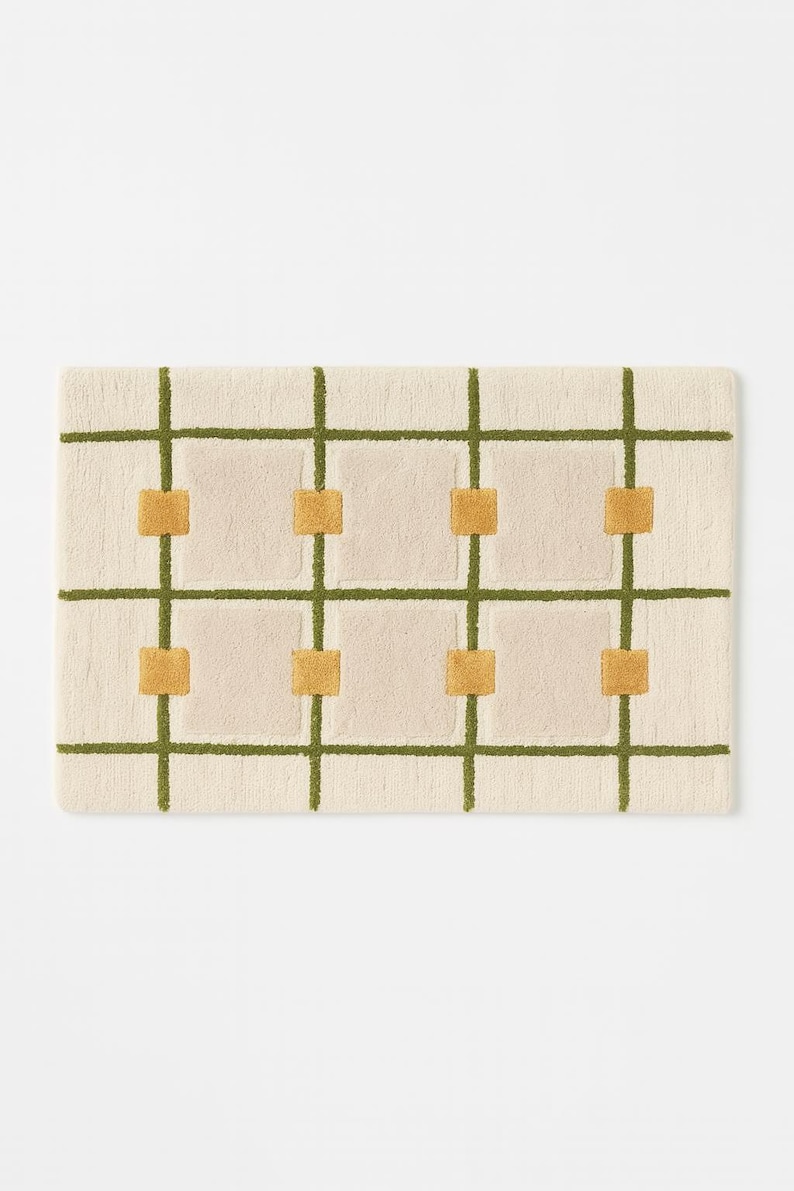 May include: Rectangular rug with a cream-colored background, featuring a grid pattern of olive green lines and squares. The squares are filled with a mustard yellow color. The rug has a soft, textured appearance.