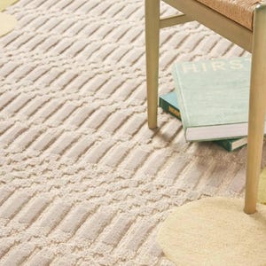 Pu&ograve; includere: Tappeto beige e crema con motivo geometrico strutturato. Il tappeto ha un bordo smerlato con dettagli gialli e verdi. Parzialmente visibile una sedia in legno con seduta intrecciata e un libro.