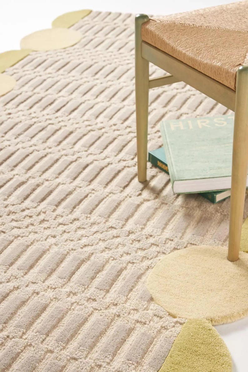 Pu&ograve; includere: Tappeto beige e crema con motivo geometrico strutturato. Il tappeto ha un bordo smerlato con dettagli gialli e verdi. Parzialmente visibile una sedia in legno con seduta intrecciata e un libro.