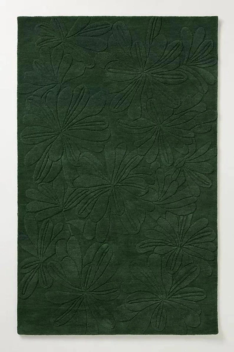 May include: A dark green rug with a repeating floral pattern. The design features large, stylized flowers with detailed leaf patterns, creating a textured, dimensional effect. The rug's color is a deep, forest green.