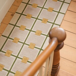 May include: A rectangular rug with a geometric design. The rug features a cream-colored background with a grid pattern of olive green lines and light gray squares. Gold-colored squares are placed within the gray squares. The rug is on a wooden floor.