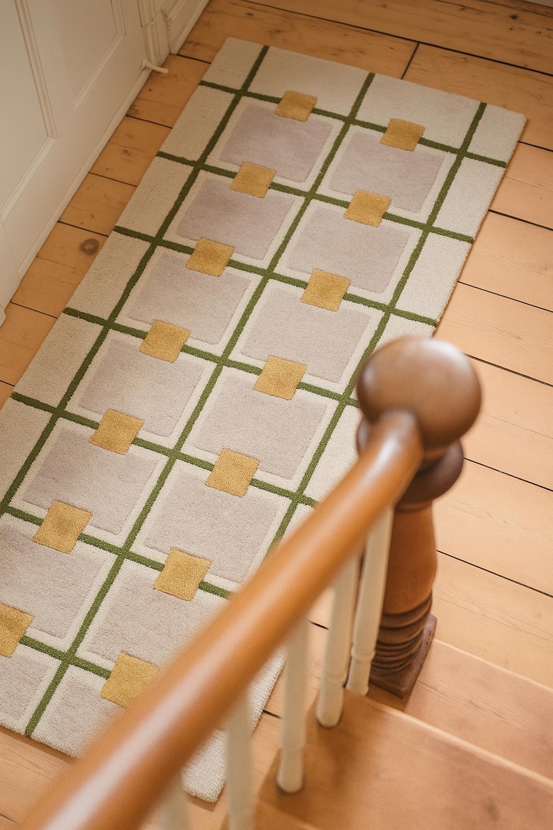 May include: A rectangular rug with a geometric design. The rug features a cream-colored background with a grid pattern of olive green lines and light gray squares. Gold-colored squares are placed within the gray squares. The rug is on a wooden floor.
