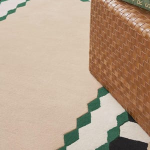 May include: Beige area rug with a geometric border in green, white, and black. A woven brown storage ottoman is partially visible on the right side, with a green book on top.