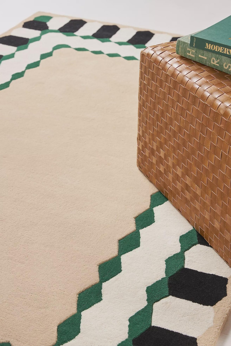 May include: Beige area rug with a geometric border in green, white, and black. A woven brown storage ottoman is partially visible on the right side, with a green book on top.