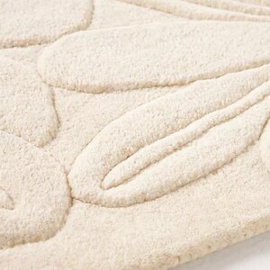 Puede incluir: Primer plano de una alfombra de ba&ntilde;o texturizada de color crema. La alfombra presenta un dise&ntilde;o floral en relieve, con formas de p&eacute;talos redondeadas y l&iacute;neas curvas. El material parece suave y afelpado, con una superficie ligeramente irregular.
