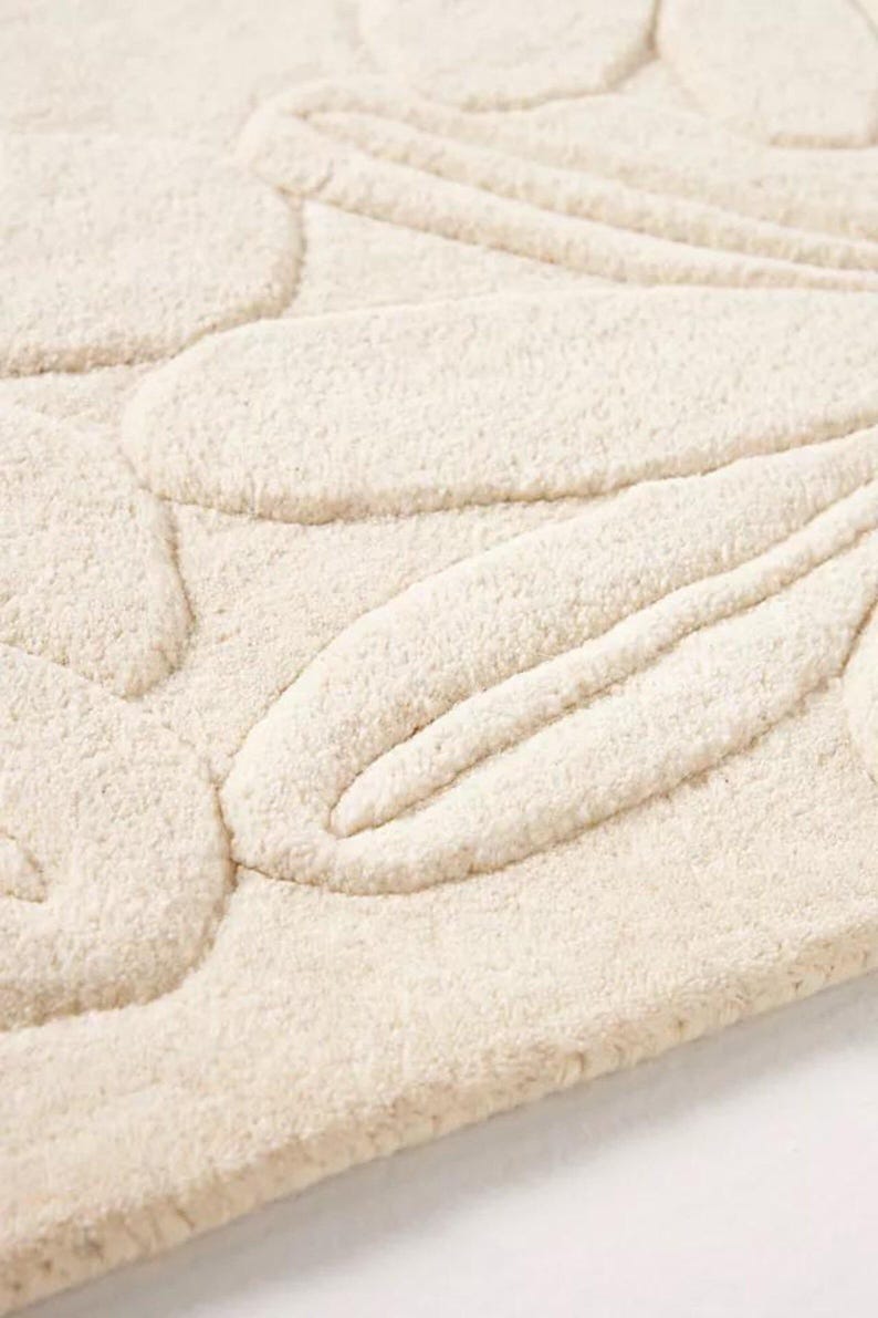 Puede incluir: Primer plano de una alfombra de ba&ntilde;o texturizada de color crema. La alfombra presenta un dise&ntilde;o floral en relieve, con formas de p&eacute;talos redondeadas y l&iacute;neas curvas. El material parece suave y afelpado, con una superficie ligeramente irregular.