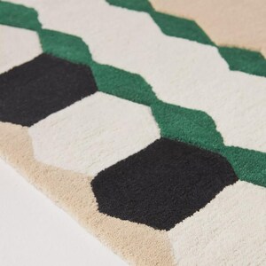 May include: Close-up of a geometric rug with a repeating pattern of black, white, green, and beige shapes. The rug has a textured surface and a modern design, suitable for home decor.