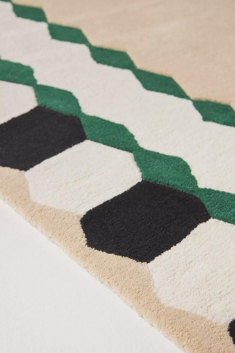 May include: Close-up of a geometric rug with a repeating pattern of black, white, green, and beige shapes. The rug has a textured surface and a modern design, suitable for home decor.