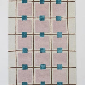 Op de afbeelding: Rechthoekig tapijt met een cr&egrave;mekleurige achtergrond en een rasterpatroon van bruine lijnen. Roze vierkanten zijn verspreid in het raster, elk met een turquoise vierkant bovenaan. Het tapijt heeft een modern, geometrisch ontwerp.