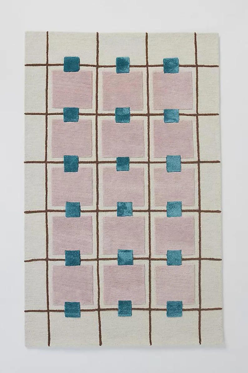 Op de afbeelding: Rechthoekig tapijt met een cr&egrave;mekleurige achtergrond en een rasterpatroon van bruine lijnen. Roze vierkanten zijn verspreid in het raster, elk met een turquoise vierkant bovenaan. Het tapijt heeft een modern, geometrisch ontwerp.