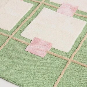 Puede incluir: Una alfombra afelpada con un dise&ntilde;o geom&eacute;trico. La alfombra presenta un fondo verde claro con cuadrados color crema y cuadrados rosas de acento. L&iacute;neas beige forman un patr&oacute;n de cuadr&iacute;cula sobre el fondo verde, creando el dise&ntilde;o geom&eacute;trico.