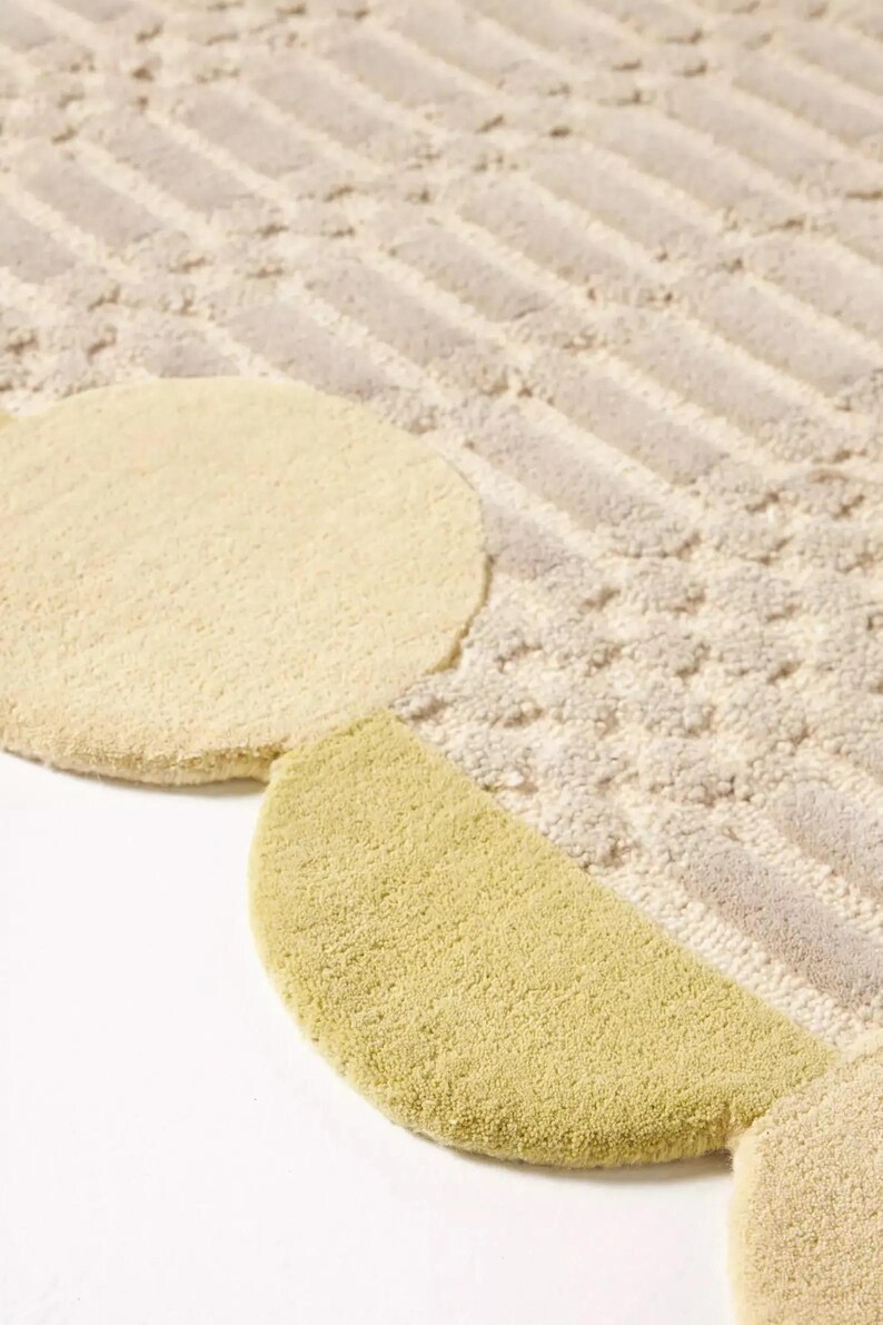Pu&ograve; includere: Primo piano di un tappeto strutturato con un design geometrico. Il tappeto presenta un motivo a strisce e cerchi beige e grigio chiaro. Una sezione del tappeto &egrave; un semicerchio verde chiaro.