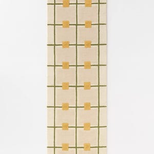 May include: Cream-colored rectangular rug with a grid pattern of olive green lines and gold squares. The rug is a long, narrow runner, suitable for hallways or entryways. The design is simple and modern, with a geometric aesthetic.