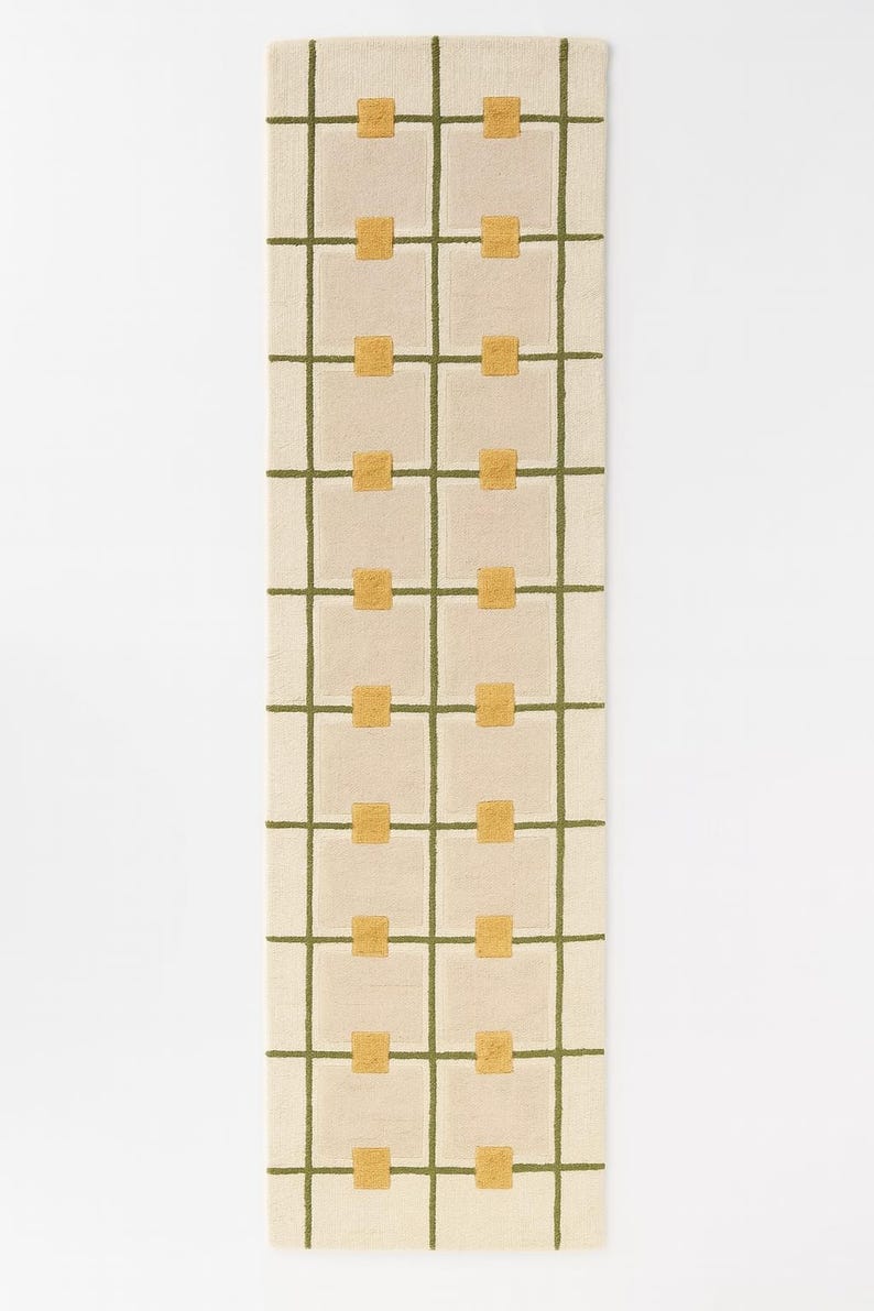 May include: Cream-colored rectangular rug with a grid pattern of olive green lines and gold squares. The rug is a long, narrow runner, suitable for hallways or entryways. The design is simple and modern, with a geometric aesthetic.